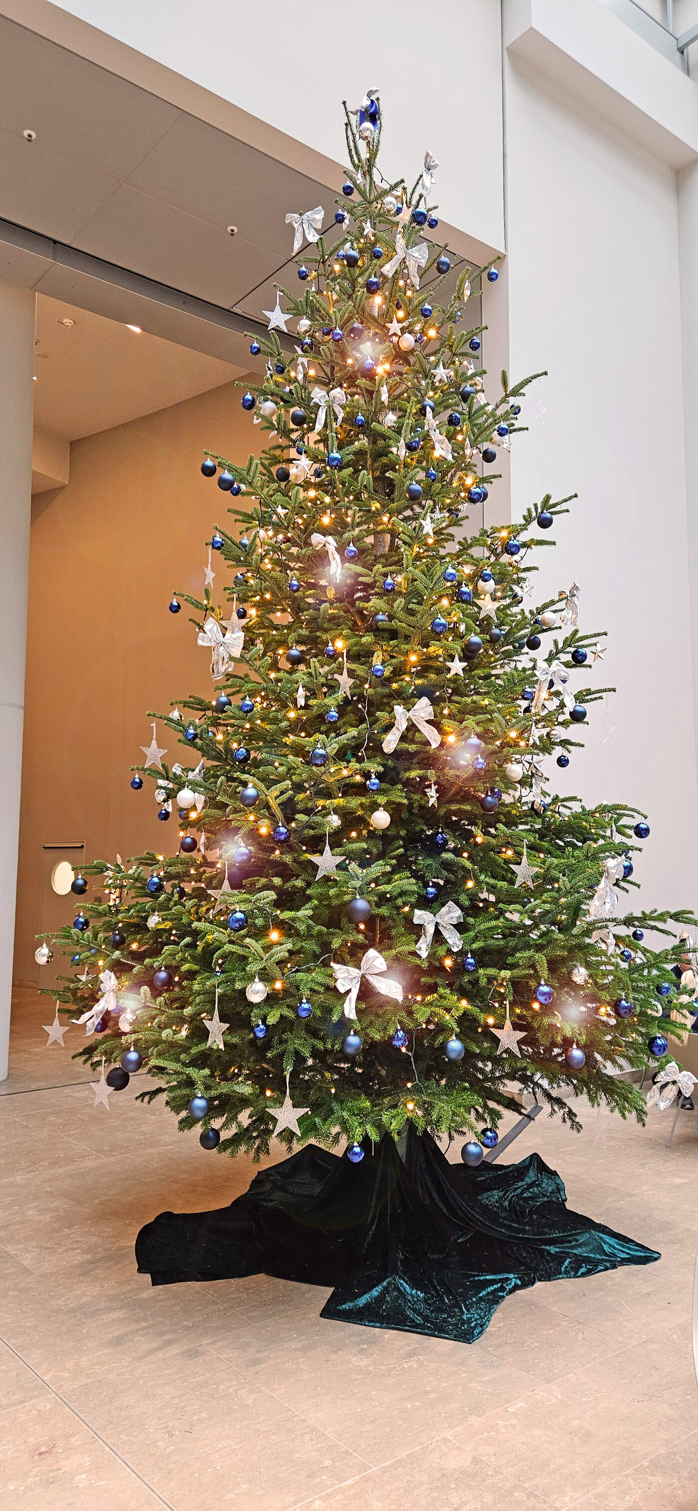 Ein festlich geschmückter Weihnachtsbaum mit silbernen Schleifen, blauen und weißen Kugeln sowie funkelnden Sternen steht in einer modernen Empfangshalle. Die warmen Lichter verleihen dem Raum eine gemütliche, festliche Atmosphäre.