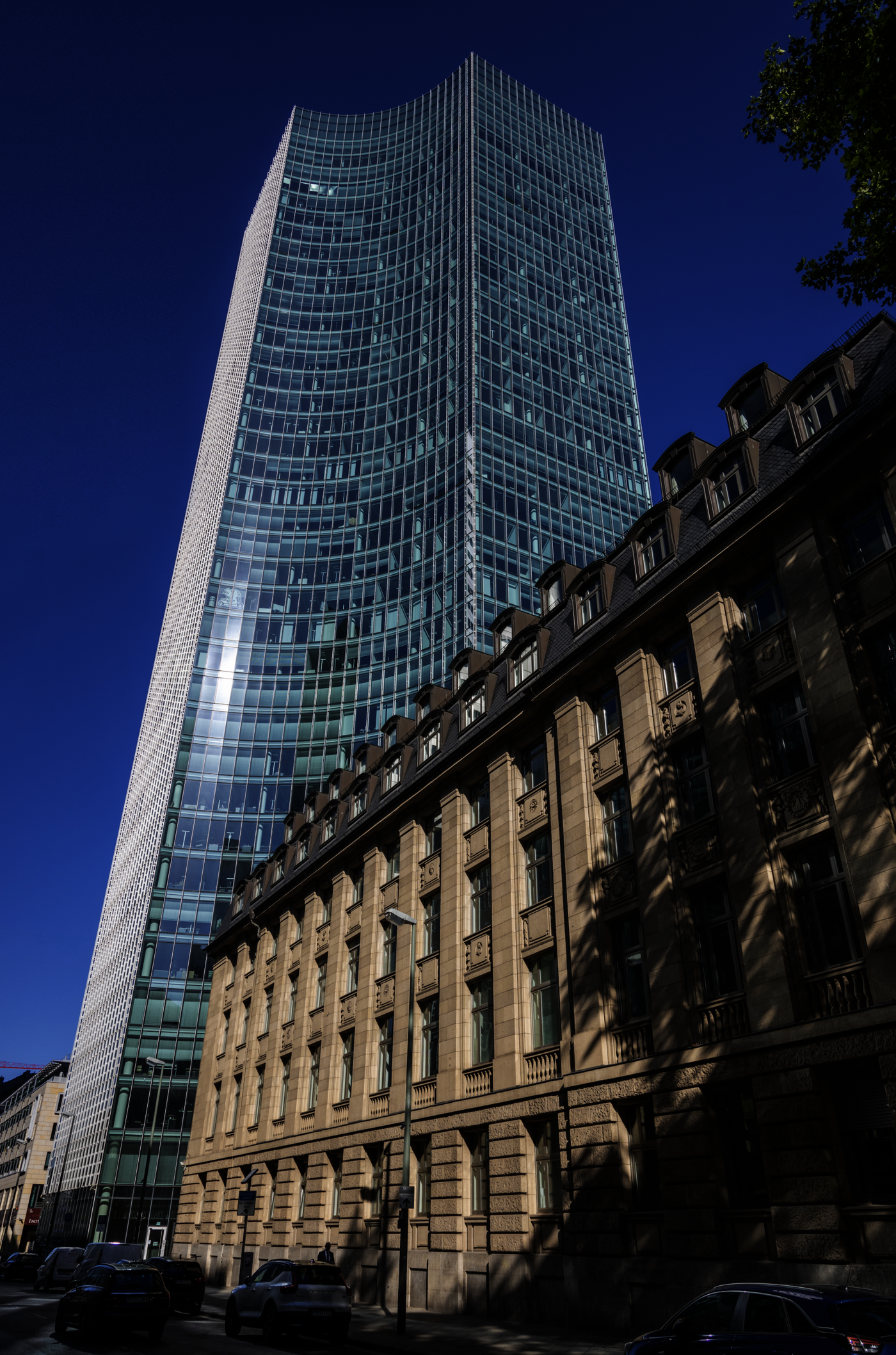 Hoher, geschwungener Glaswolkenkratzer hinter einem niedrigeren historischen Sandsteingebäude in einer Innenstadtstraße, bei blauem Himmel und mit parkenden Autos im Vordergrund.
