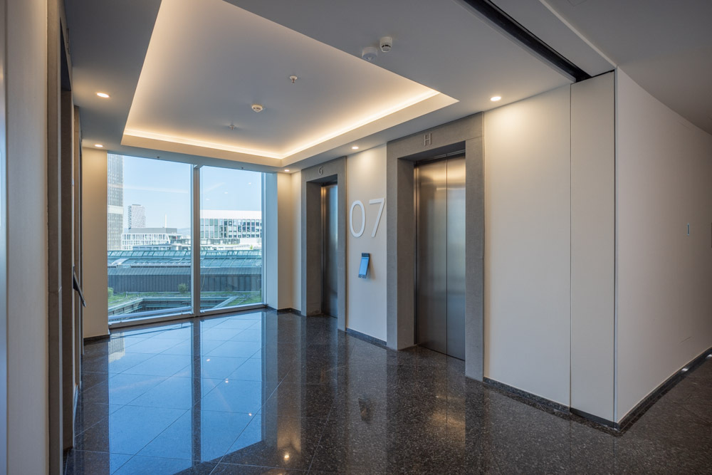 Bright elevator lobby at SKYPER Frankfurt with modern elevators, stone flooring, floor signage and views of Frankfurt city center.